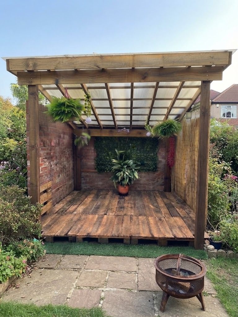 Pérgola de madera techada con policarbonato y piso de palets, con muro verde y helechos colgantes
