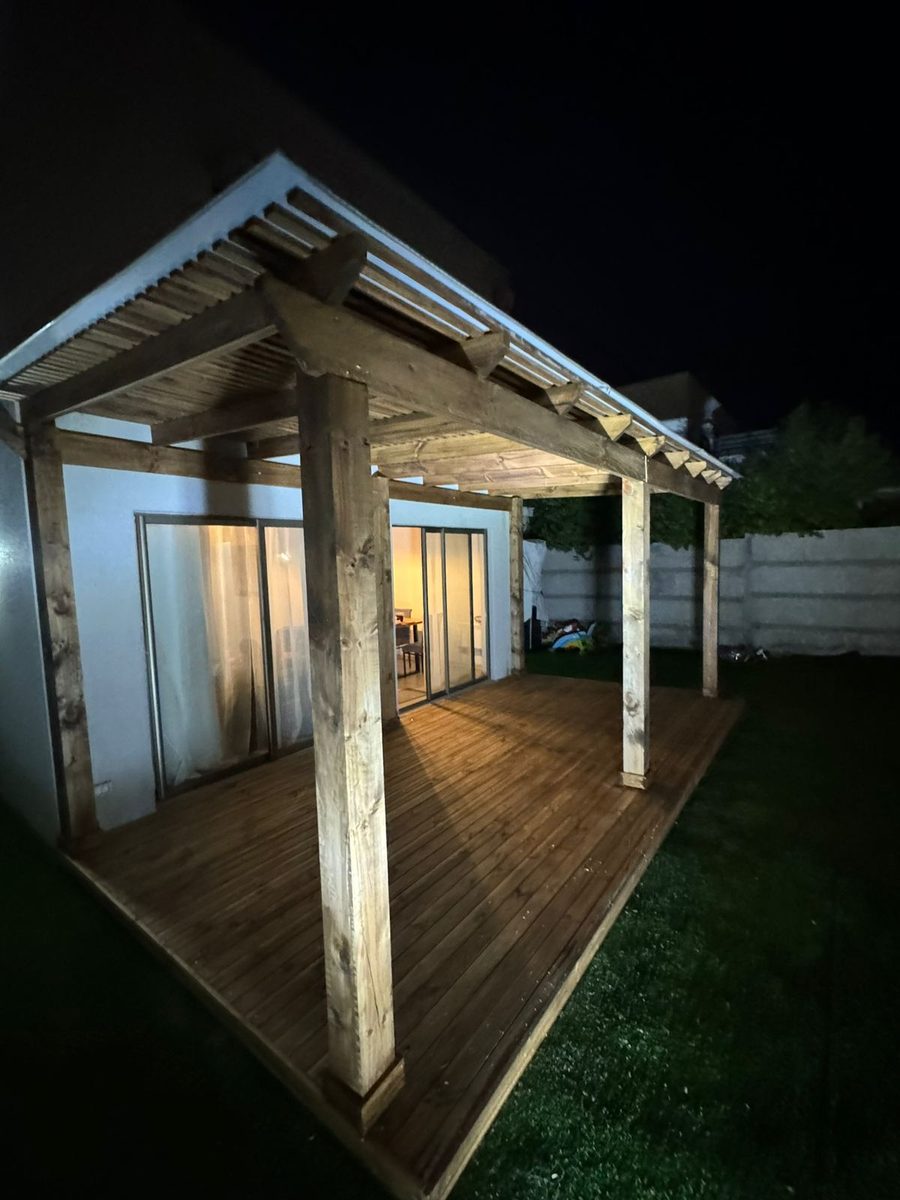 Deck de madera bajo una pérgola iluminada de noche, junto a la casa